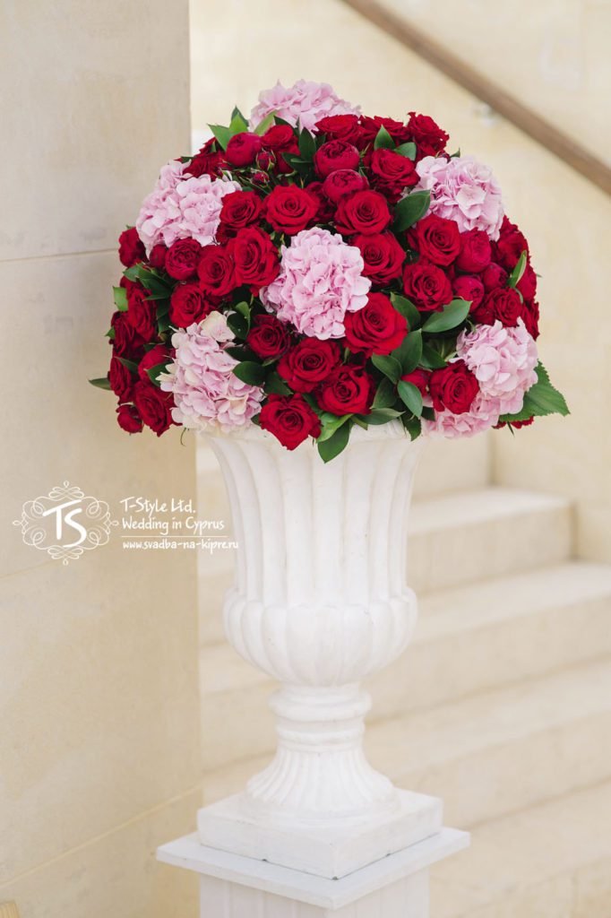 Большая белая амфора на подставке с композицией из красных роз, пионовидных роз и гортензии розового цвета с небольшим добавлением зелени